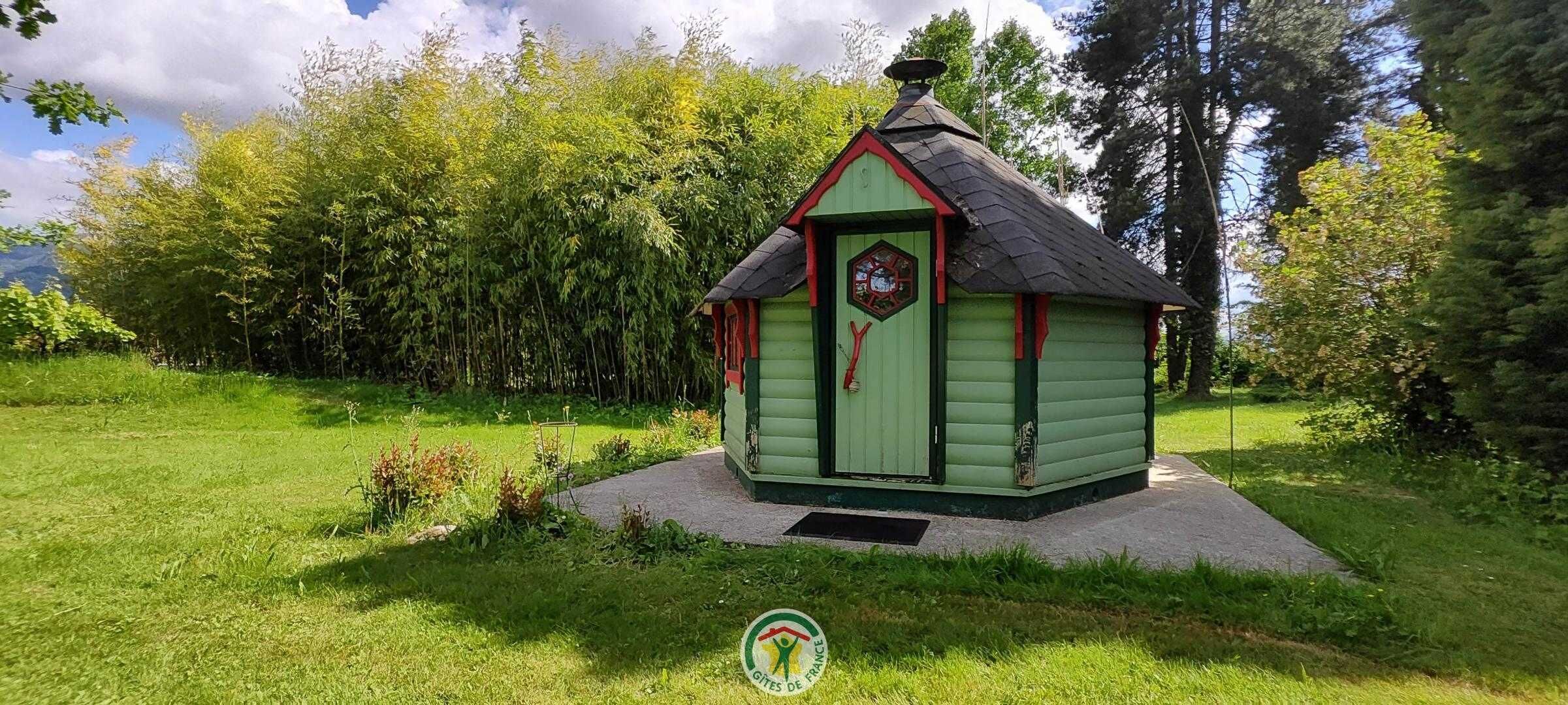 A small, single-story wooden structure with a green door and a red trim, surrounded by lush greenery and trees.