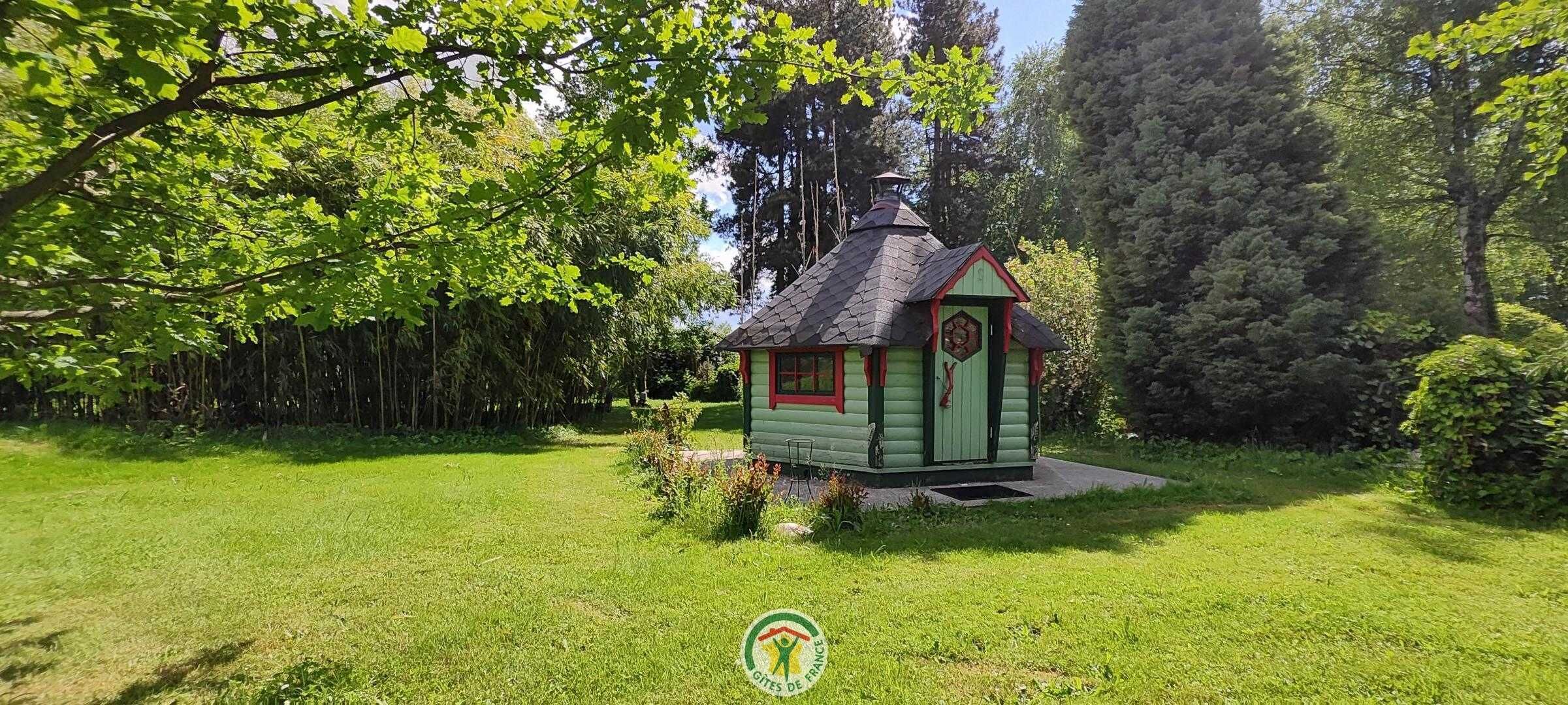 A small, green wooden structure with a red roof, surrounded by lush greenery and trees.