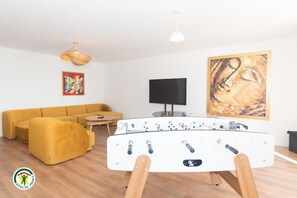 A living room with a sofa, a foosball table, a television, and artwork on the walls. - Domaine des Étoiles - La Villa Vacation Rental (GILLONNAY)