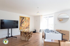 A modern living room with a flat-screen TV, a wooden coffee table, a foosball table, and a large abstract painting on the wall. - Domaine des Étoiles - La Villa Vacation Rental (GILLONNAY)