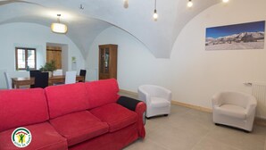 A living room with a red sofa, white armchairs, a dining area with a table and chairs, a cabinet, and a painting on the wall.