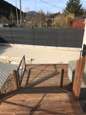Una terraza de madera con baranda metálica, un muro de concreto y un vehículo al fondo.