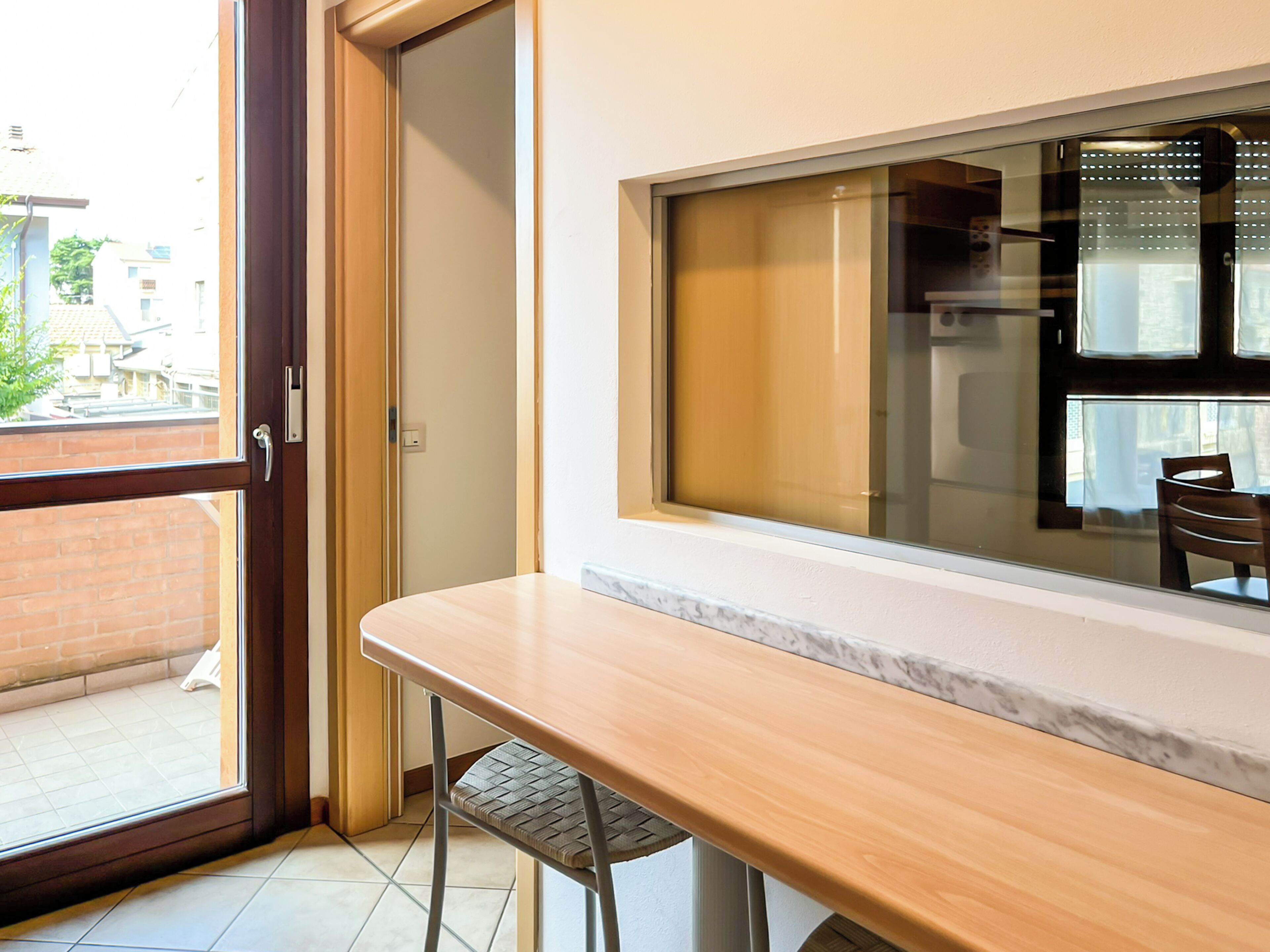 Una stanza con una porta-finestra in vetro scorrevole che si apre su uno spazio esterno, un tavolo di legno con una sedia e un'ampia finestra con persiane.