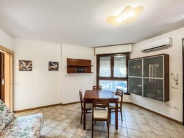 A dining area with a wooden table and chairs, a window with blinds, a wall-mounted air conditioner, and a small shelf with books.