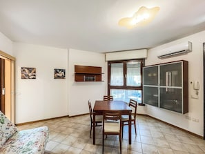 Un comedor con una mesa y sillas de madera, una ventana con persianas, un aire acondicionado montado en la pared y un estante pequeño con libros.