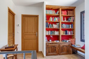Une pièce avec une bibliothèque en bois remplie de livres, une petite table sur laquelle repose un livre et un banc agrémenté d’un coussin.