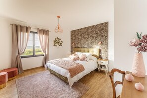 Ein Schlafzimmer mit einem großen Bett, einem Nachttisch, einem Schreibtisch, einem Stuhl, einem Fenster mit Vorhängen und einer Wand mit einem Blumenmuster.