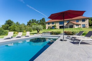 Une piscine entourée de chaises longues et agrémentée d’un parasol rouge, le tout niché au cœur d’un jardin aménagé, avec une maison à deux étages en arrière-plan.