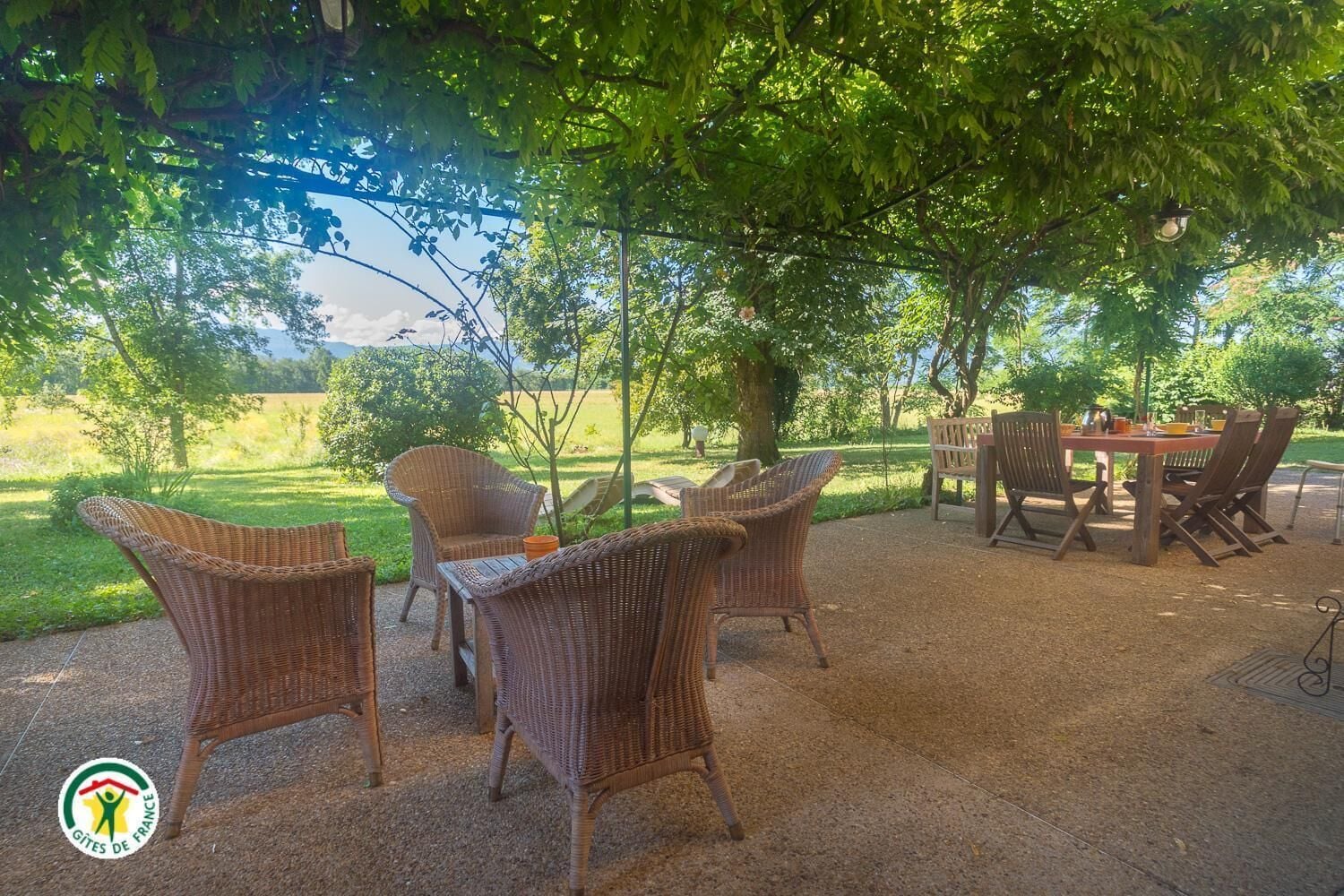Un patio al aire libre con mobiliario de mimbre y una mesa, rodeado de árboles y un campo cubierto de césped.