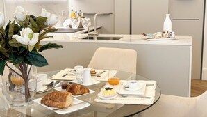 Un espace repas moderne avec une table en verre, des chaises blanches et une variété d’aliments pour le petit-déjeuner, notamment des croissants, des viennoiseries et un panier de fruits.