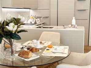 Un comedor moderno con una mesa de vidrio, sillas blancas y una selección de productos para el desayuno, como cruasanes, pasteles y un bol de fruta.