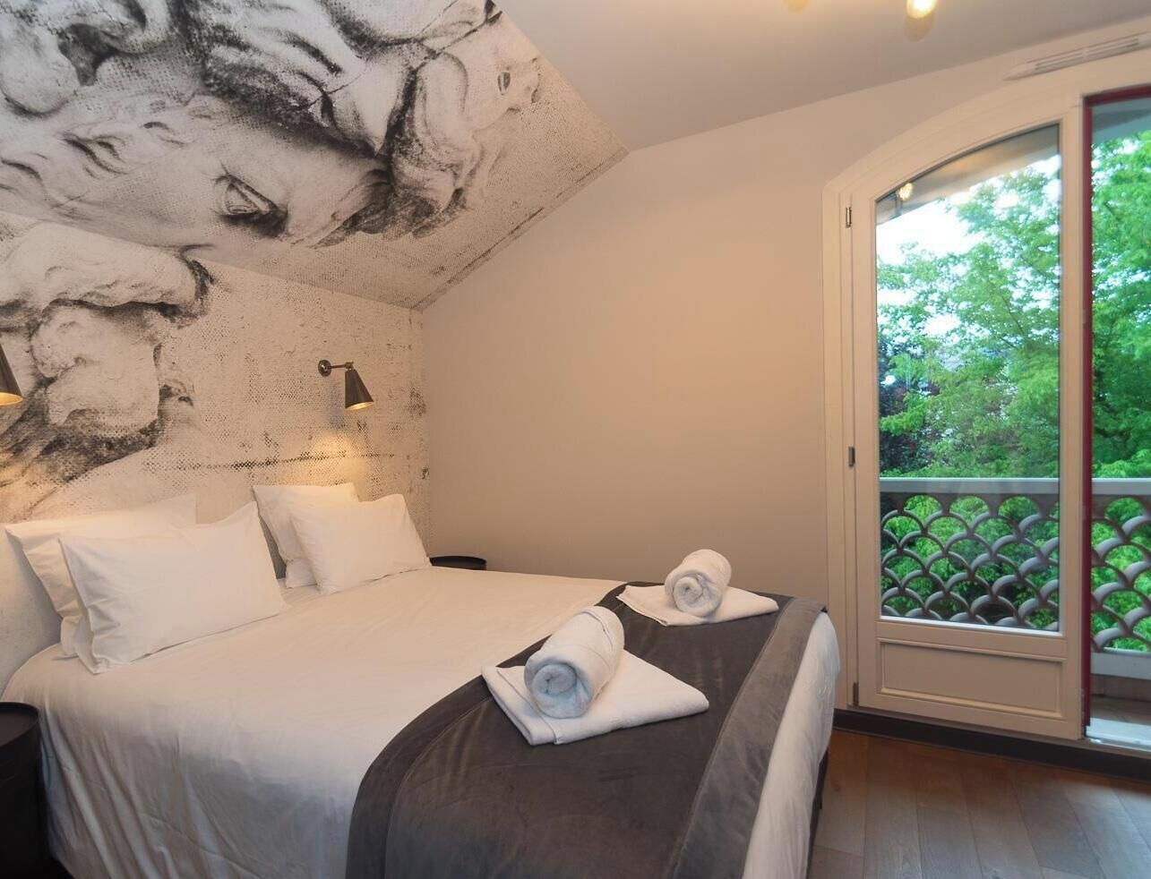 A hotel room with a bed, two pillows, a nightstand, a wall mural, a window with a view of greenery, and a balcony with a lattice railing.