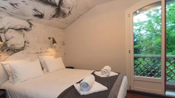 A hotel room with a bed, two pillows, a nightstand, a wall mural, a window with a view of greenery, and a balcony with a lattice railing.