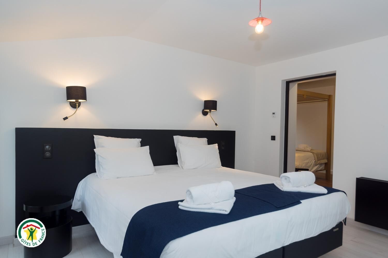 A modern hotel room with a large bed, two bedside lamps, a nightstand, and a visible door leading to another room.