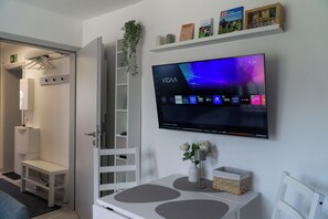 A modern hotel room with a flat-screen TV mounted on the wall, a white console table with decorative items, and a built-in shelving unit with books and a plant.