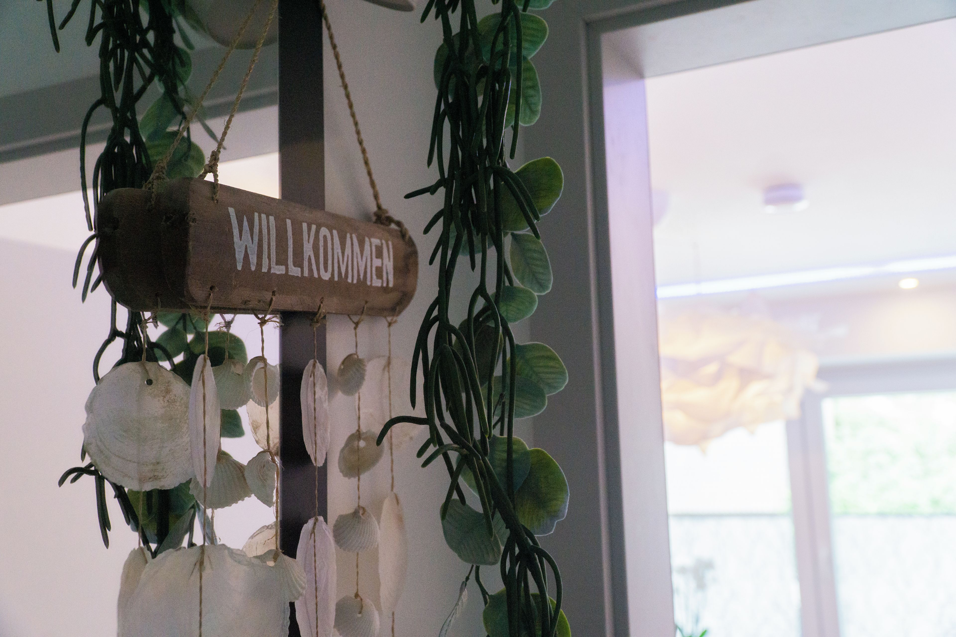 Ein hölzernes Schild mit der Aufschrift 'WILLKOMMEN', das an der Decke hängt, umgeben von grünem Laub und weißen Blüten.
