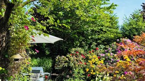 En have med forskellige planter og blomster, et terrassemiljø med et bord og stole, og et træ der giver skygge.
