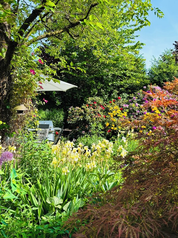 A garden with a variety of plants and flowers, a patio area with a table and chairs, and a tree providing shade.