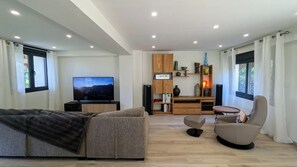A modern living room with a flat-screen TV, a sofa, a chair, a wooden bookshelf, and a dining area with a table and chairs. - From Combeloup - MURIANETTE (MURIANETTE)