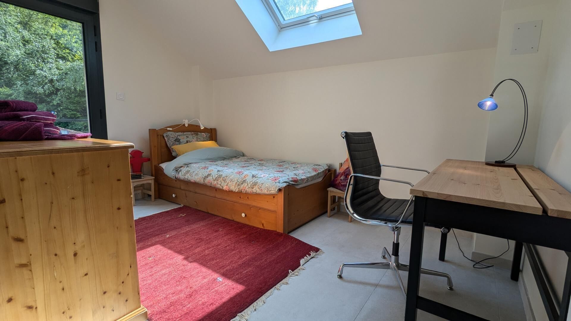 Ein Schlafzimmer mit Oberlicht, einem Bett, einem Schreibtisch, einem Stuhl und einem roten Teppich.