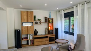 A modern living room with a wooden entertainment unit, a round wicker table, a sofa, and a view through a window with curtains. - From Combeloup - MURIANETTE (MURIANETTE)