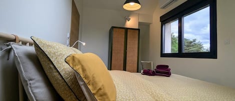 Un dormitorio con cama, almohadas, una lámpara de noche, una ventana y un equipo de aire acondicionado.