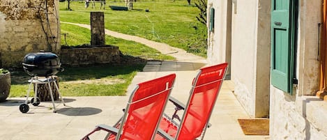 A patio with two red chairs, a barbecue grill, and a view of a green field with a cow.