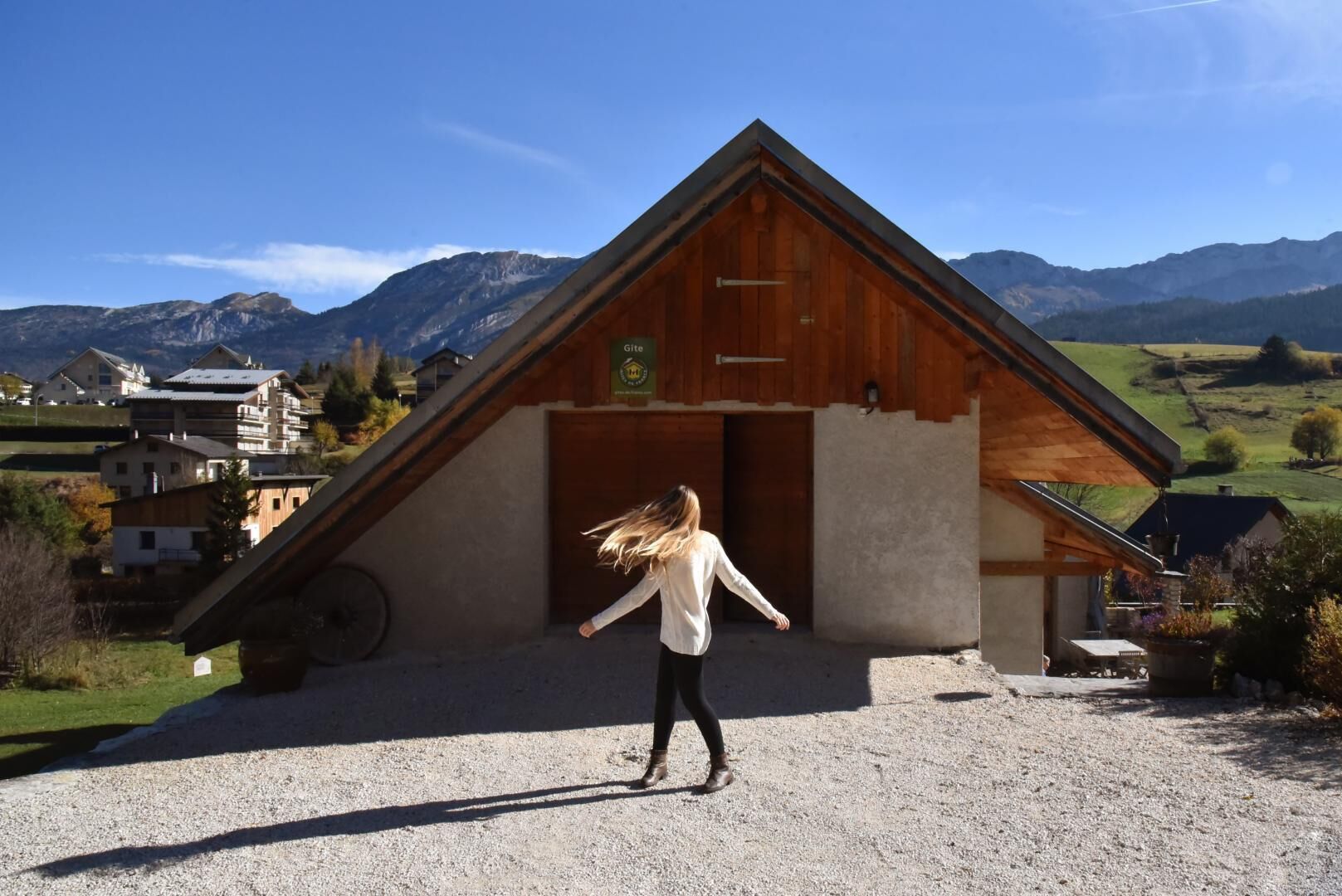 Une personne se tient devant un bâtiment en bois doté d’un toit en pente, avec en arrière-plan des montagnes et des maisons.