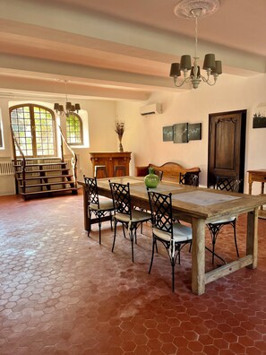 Amplia habitación con una gran mesa de comedor de madera, suelo con patrón hexagonal y una escalera con vistas a la vegetación exterior.
