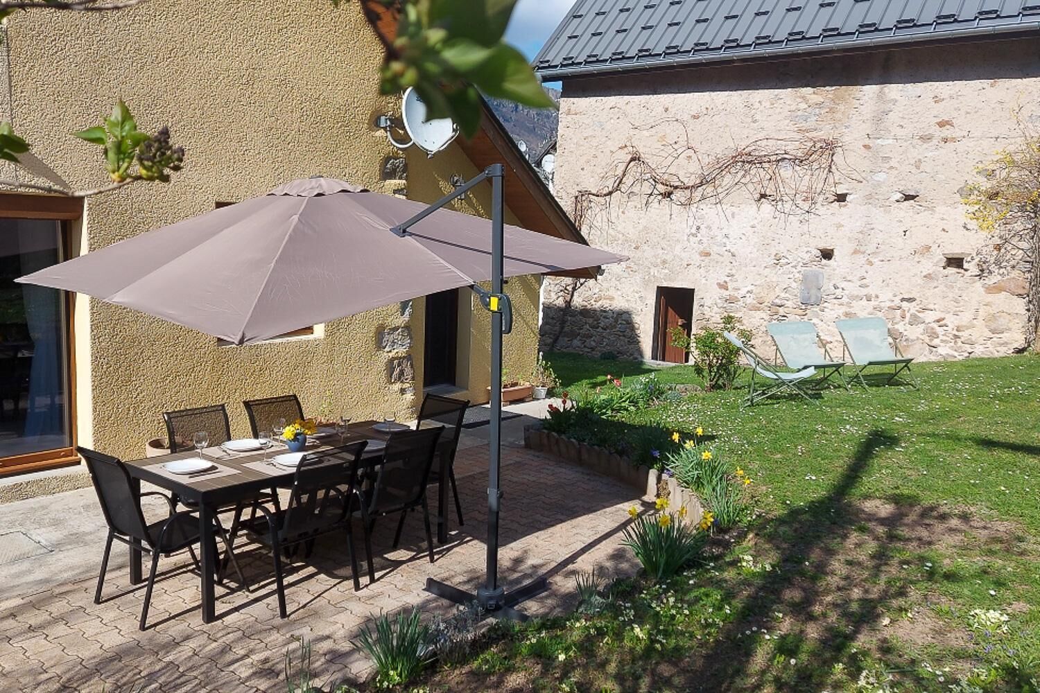 Eine Terrasses mit Tisch und Stühlen, einem großen Sonnenschirm und einem Steinhaus im Hintergrund.