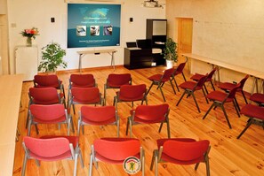 A conference room with red chairs arranged in rows, a projector screen displaying a presentation, and a small stage area with a laptop. - The Plantier Estate Guesthouse (SAINT HILAIRE DE LA COTE)