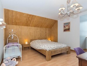 Un dormitorio con techo de madera, una cama con colcha floral, una mesita de noche con lámpara, una silla de madera y un cuadro colgado en la pared.