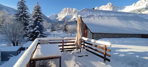 A wooden bench with snow accumulation, a metal-framed table, and a building with a corrugated metal roof in a snowy mountainous area. - The Violets of Grand Veymont (GRESSE EN VERCORS)