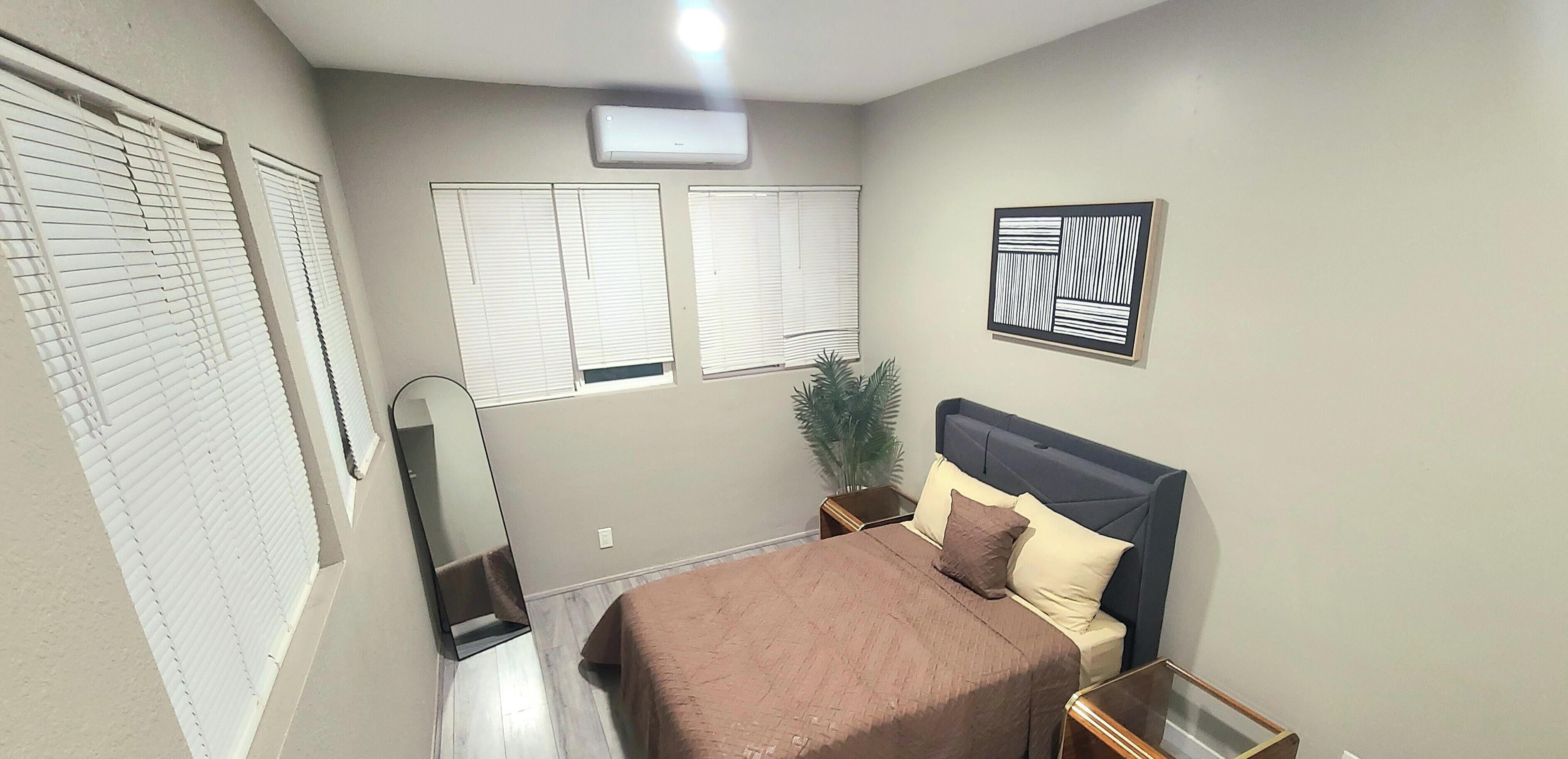 Une chambre à coucher comprenant un lit, des tables de chevet, un miroir, une plante en pot et un climatiseur fixé au mur.