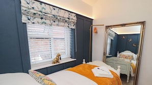 A bedroom with a bed, a window with blinds, a mirror, and a small dog figurine on the windowsill. - Acorn Cottage (Yarm)