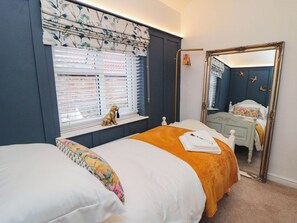 A bedroom with a bed, a window with blinds, a mirror, and a small dog figurine on the windowsill. - Acorn Cottage (Yarm)