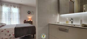 A hotel room with a bed, a bedside table with a lamp, a bathroom with a sink, and a mirror. - Gite de la Manonière - 4**** (SAINT DIDIER DE LA TOUR)