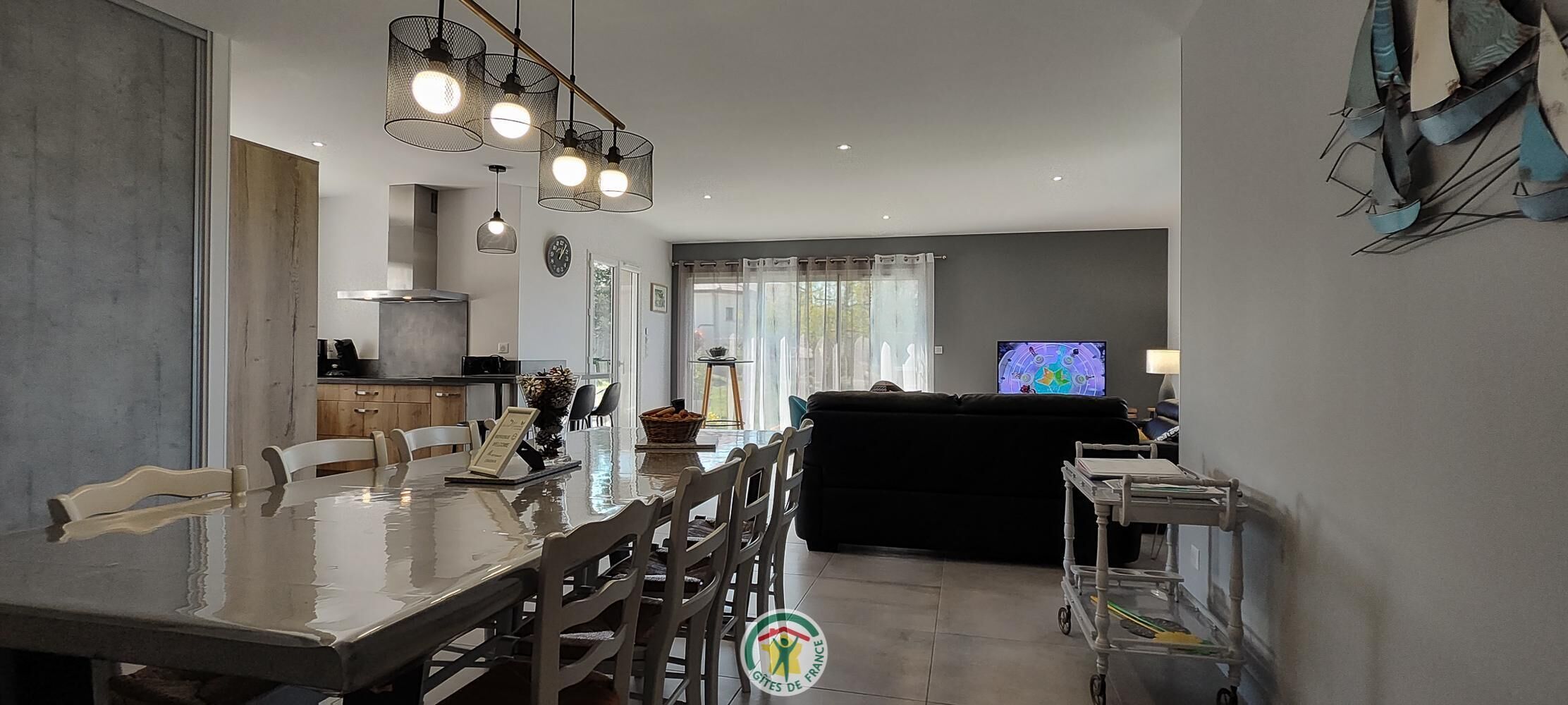 A modern kitchen with a dining area, a living room with a black sofa, and a television.