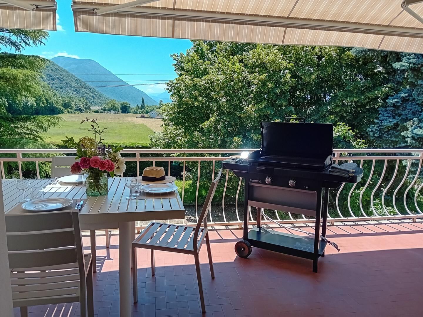 An outdoor dining area with a table set for two, a grill, and a view of a green landscape with mountains in the background.