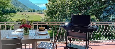 An outdoor dining area with a table set for two, a grill, and a view of a green landscape with mountains in the background.