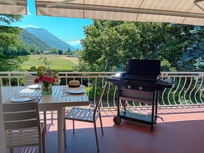 An outdoor dining area with a table set for two, a grill, and a view of a green landscape with mountains in the background.