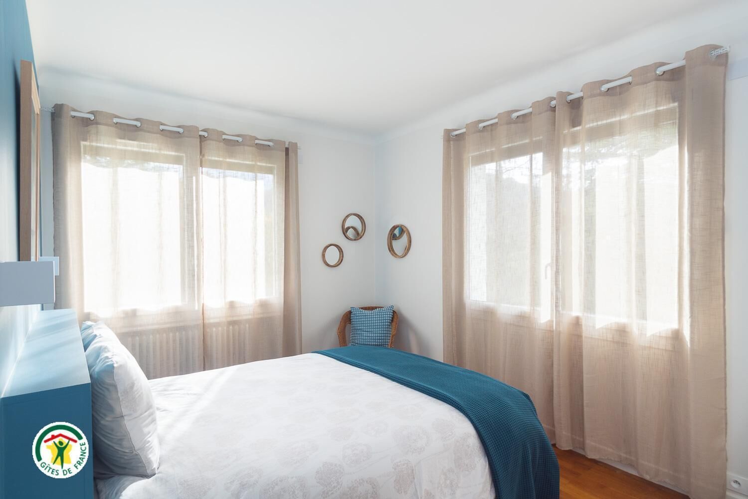 Ein Schlafzimmer mit einem Bett, zwei Fenstern mit durchsichtigen Vorhängen, einem Sessel und Wanddekorationen.