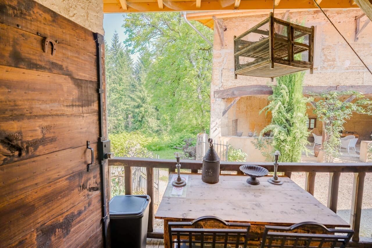 A wooden balcony with a table and chairs, overlooking a garden and a building with stone walls.