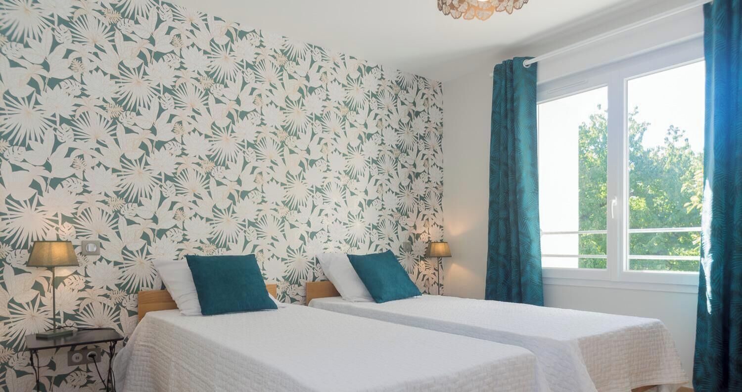 Una habitación con dos camas, un ventanal con cortinas y una pared con estampado floral.