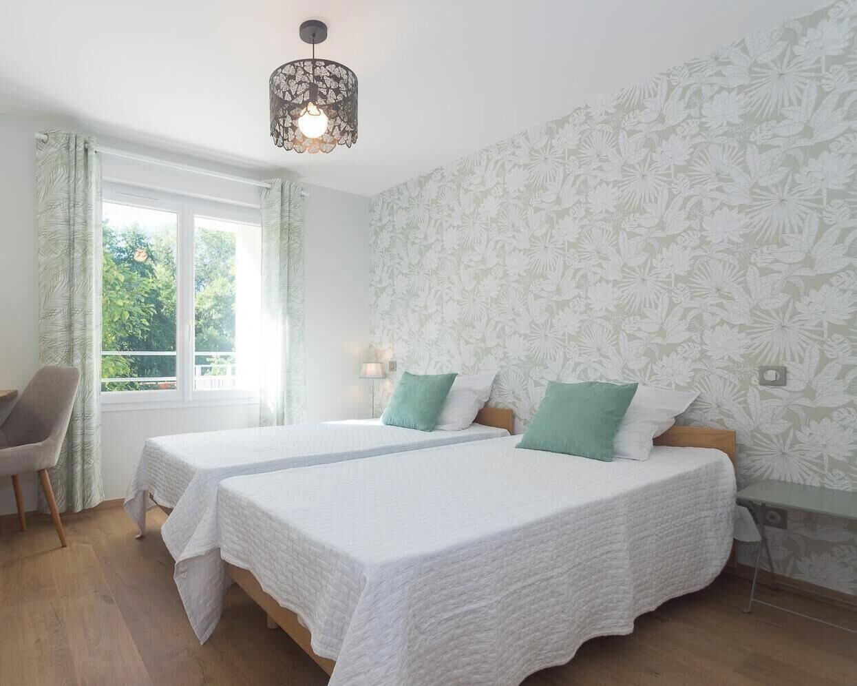 Ein Zimmer mit zwei Betten, einem Sessel, einem Fenster und einer Wand mit Blumenmuster.