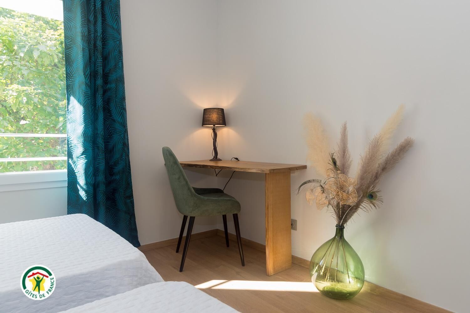 Une chambre à coucher avec un lit, un bureau avec une chaise, une lampe et un vase contenant des plantes séchées.