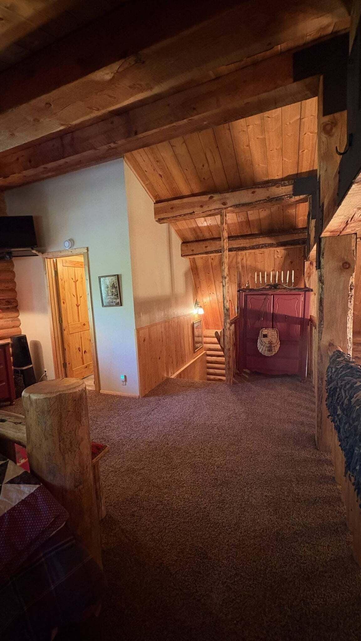 Interno di una baita in legno con una scala che conduce a una zona mansardata, un camino e una porta di legno.