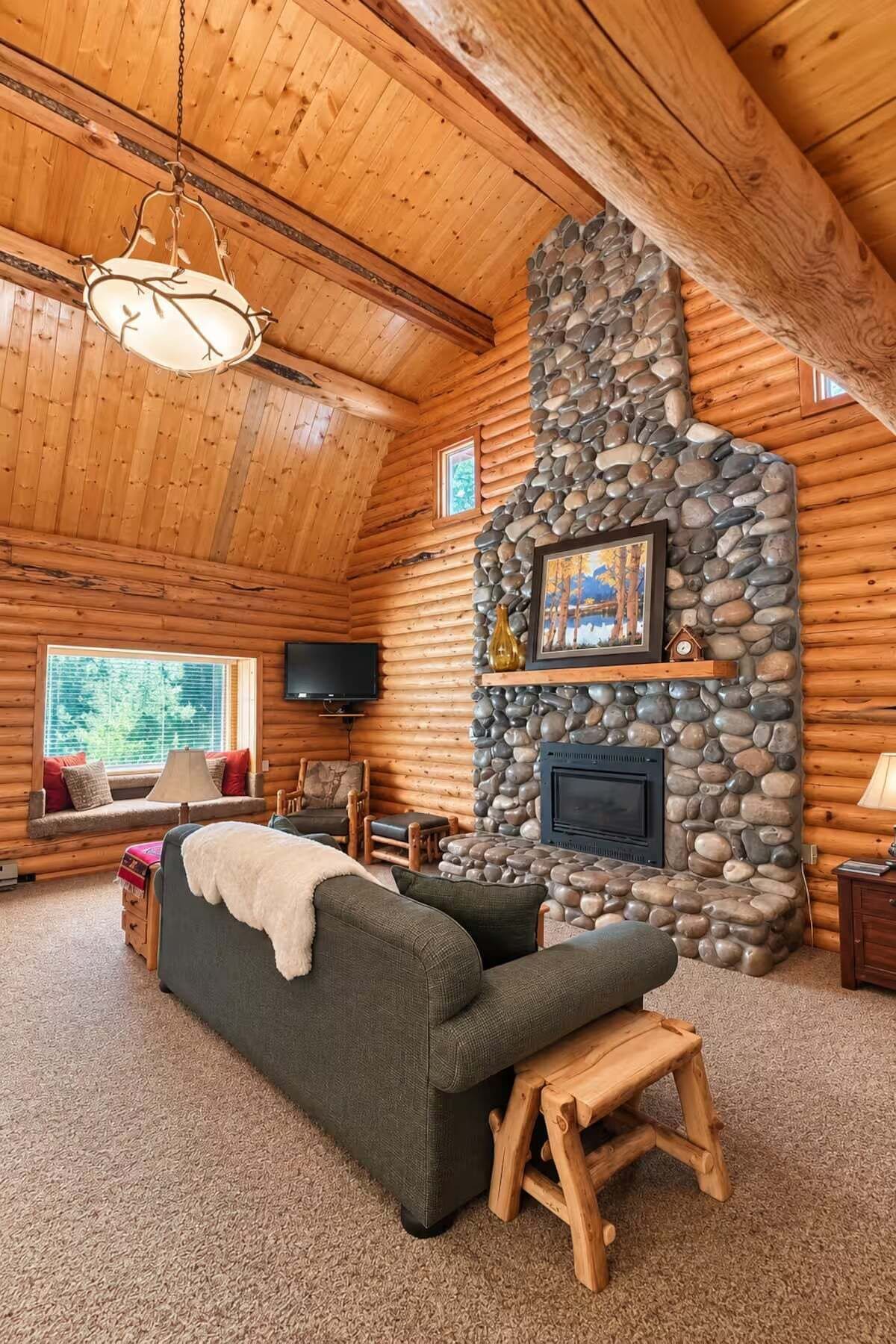 A log cabin interior with a stone fireplace, a sofa, a television, and a window with a view of trees.