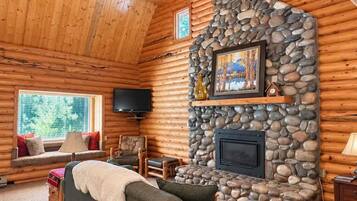A log cabin interior with a stone fireplace, a sofa, a television, and a window with a view of trees.