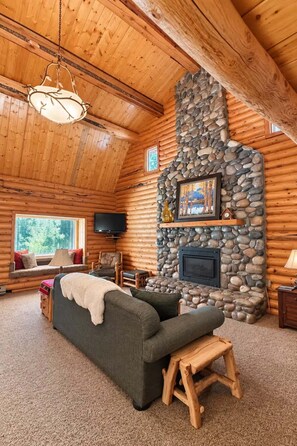 L’intérieur d’une cabane en rondins, avec une cheminée en pierre, un canapé, un téléviseur et une fenêtre donnant sur la forêt.